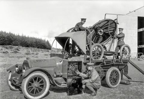 1909 Ford Model T - Three men in military-style uniforms work on a large, complex apparatus mounted on a truck chassis. The apparatus features a large paraboli…