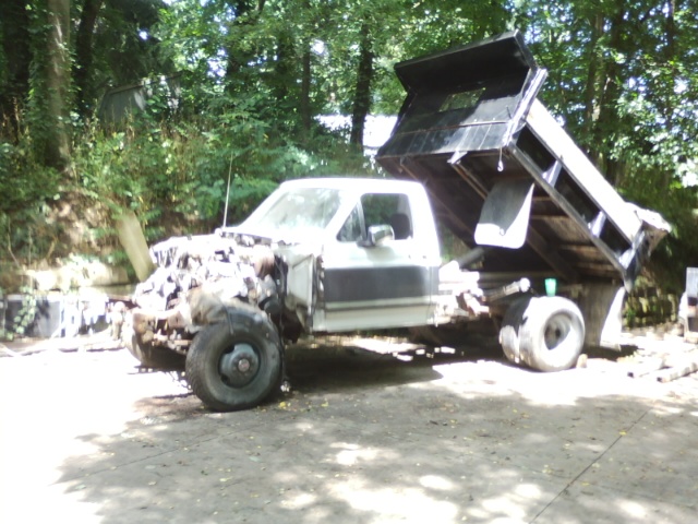 White dump truck with raised bed and damaged front end in a wooded area.
