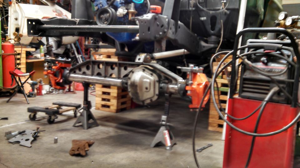 Custom front axle assembly with orange brake calipers on jack stands in a workshop.