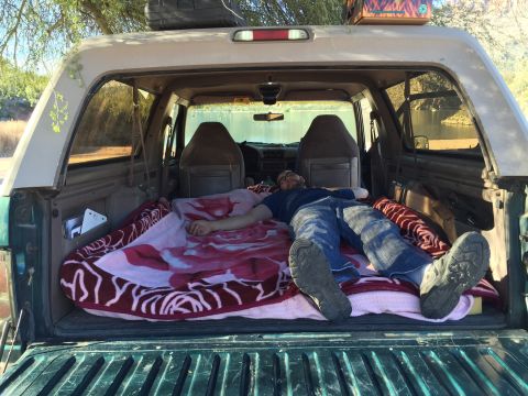 1996 Ford Bronco - A man sleeps on a blanket in the cargo area of a Ford Bronco, which is parked outdoors near a body of water.