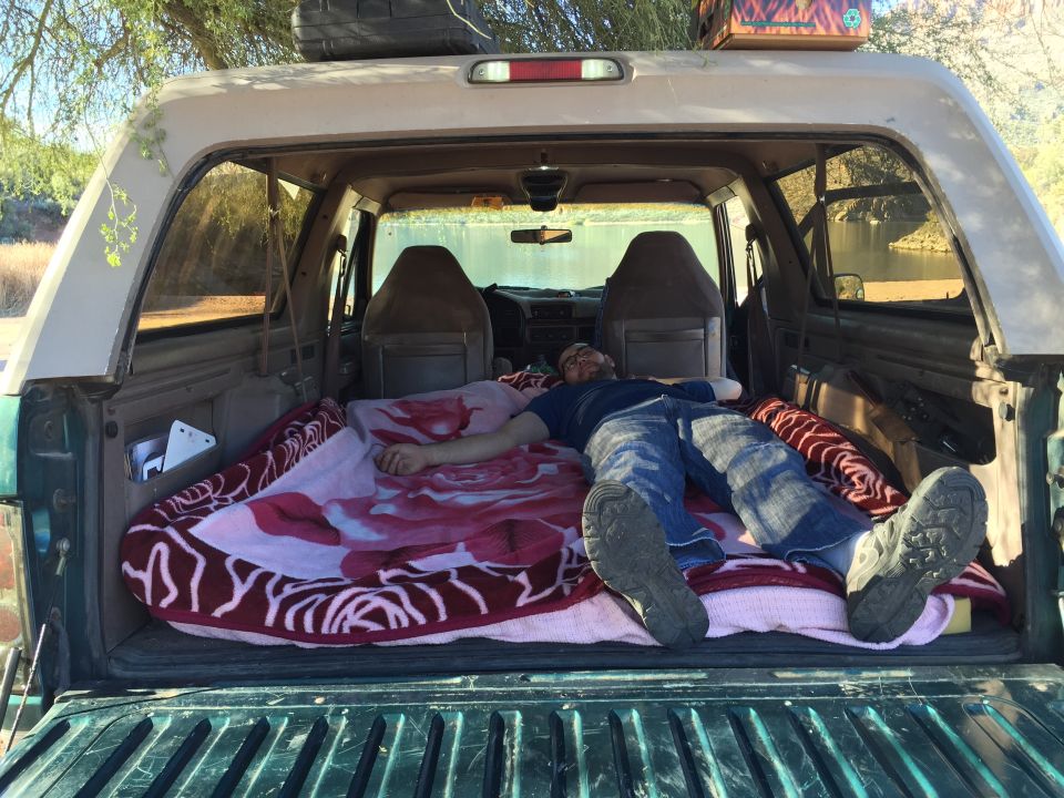 Man sleeping in the back of a Ford Bronco with a lake in the background.