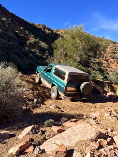 1996 Ford Bronco - A teal and tan 1996 Ford Bronco is shown from the rear quarter angle, driving on a dirt path through a rocky, arid landscape.