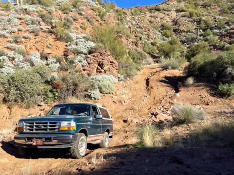 1996 Ford Bronco - A dark green 1996 Ford Bronco is parked on a dirt trail in a desert landscape with a steep, rocky hillside behind it.