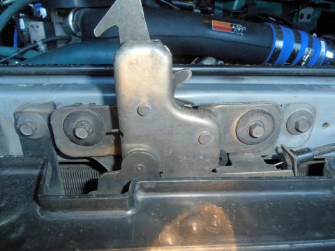 1990 Ford Bronco - A close-up view of the hood latch mechanism on a vehicle, showing metal and plastic components secured by bolts and screws. A black K&N air…