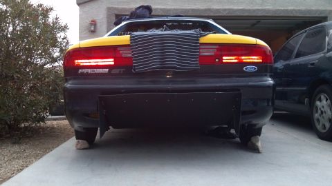 1995 Ford Probe - The rear view of a yellow and black Ford Probe GT is shown, parked in a driveway with a dark sedan to its right. The car has a custom black…