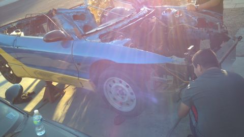 1995 Ford Probe - A blue and yellow two-door car is elevated on jack stands with its hood and front end disassembled. Two people are working on the car's eng…