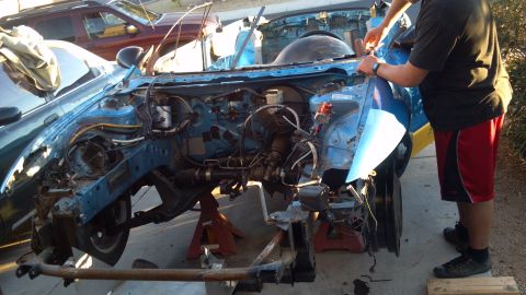 1995 Ford Probe - A blue car chassis is elevated on jack stands with its engine bay exposed. A person in red shorts and a dark shirt is working on the vehicl…