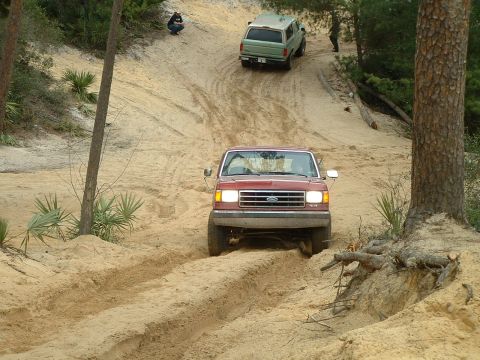 1995 Ford Bronco - Two SUVs navigate a sandy incline, with the red Ford Bronco leading the way up the challenging terrain.