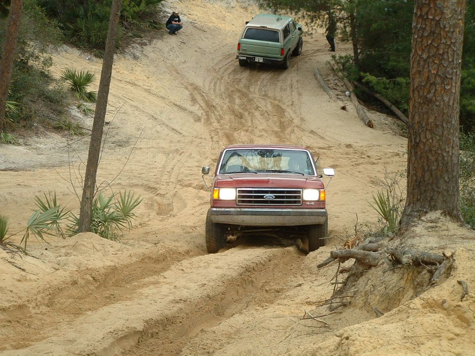 Red 1995 Ford Bronco driving up a sandy hill, with a green SUV behind it