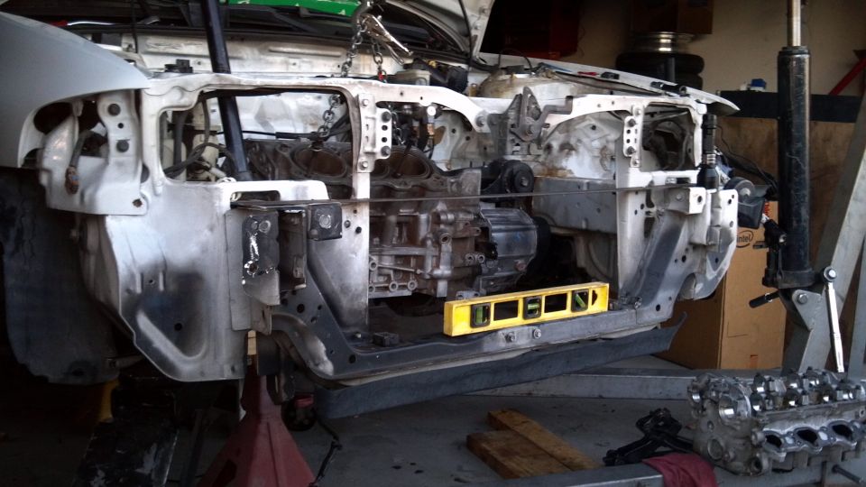 Disassembled white car front end with engine removed, cylinder head in foreground.