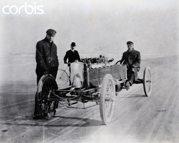 1906 Ford Model T on a sandy surface with three men, one driving