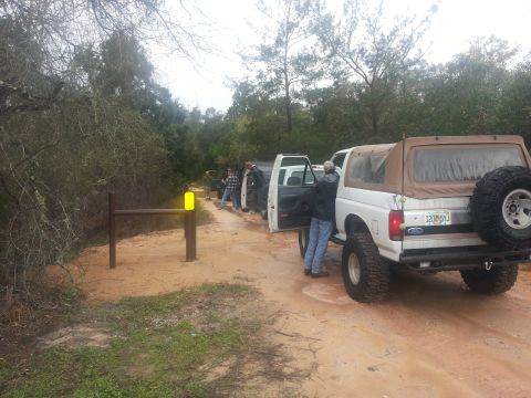 1994 Ford Bronco - Several people stand around white SUVs on a muddy dirt road with trees in the background.