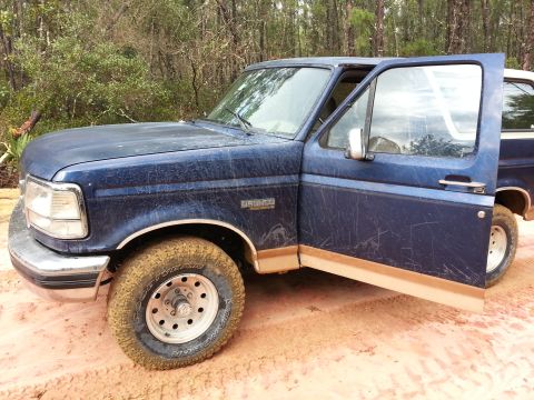 1994 Ford Bronco - A blue Ford Bronco sits with its driver's door open, surrounded by trees and dirt.