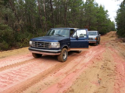 1994 Ford Bronco - Two trucks drive down a dirt road in a wooded area.
