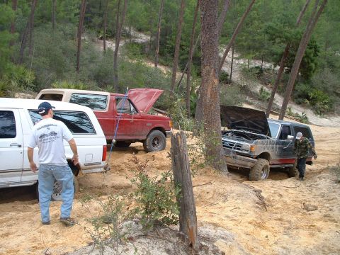 1995 Ford Bronco - Several off-road vehicles appear to be stuck in a sandy, wooded area with their hoods open.