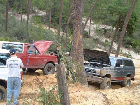 1995 Ford Bronco - Two Ford Broncos are stuck in the sand off-road, with their hoods open.