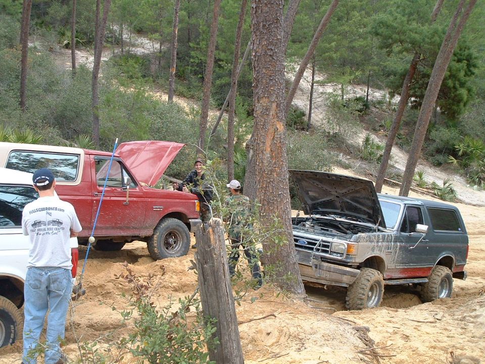 Dark green Ford Bronco with hood open, stuck in sand, next to a red Ford Bronco
