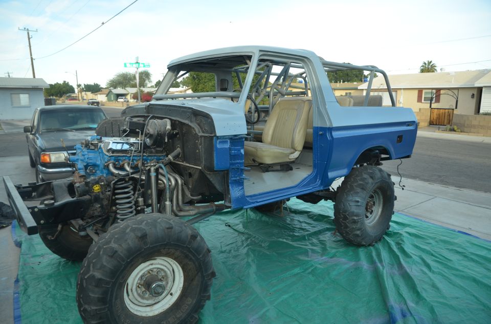 Blue and gray Ford Bronco body with exposed engine and roll cage, sitting on a green tarp. A black pickup truck is parked behind it