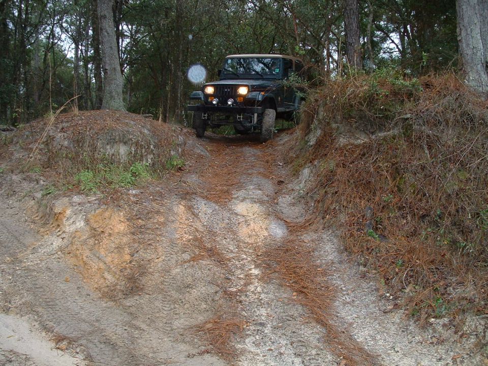 Jeep Wrangler driving up a sandy, wooded trail. Round headlights and grille indicate a TJ model