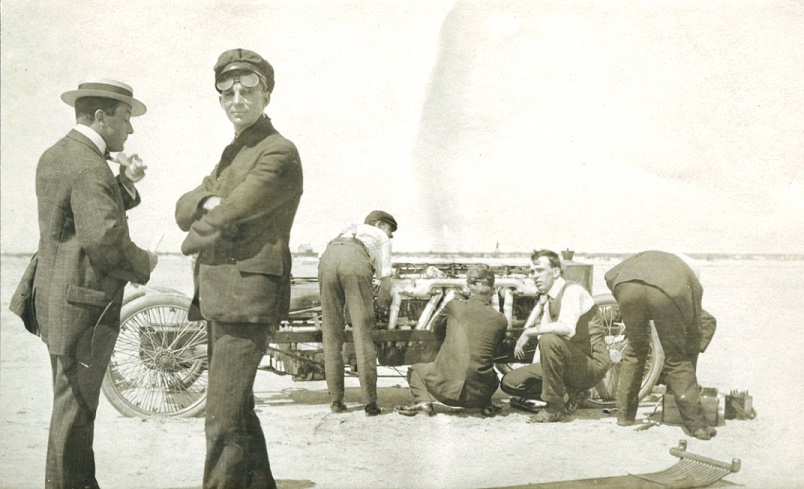 Two men stand in foreground, one talking, other looking at camera. Behind them, a group works on a large engine
