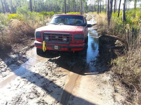 1995 Ford Bronco - A red 1995 Ford Bronco navigates a muddy path surrounded by trees and vegetation.