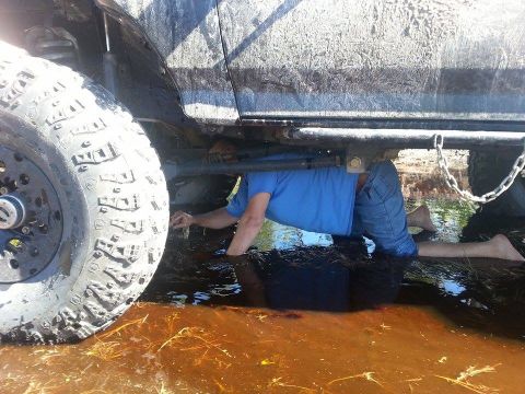 1995 Ford Bronco - A person is working under a muddy vehicle stuck in murky water.