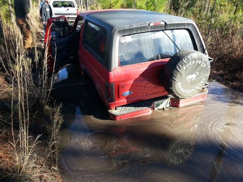 1995 Ford Bronco - A red Ford Bronco is stuck in a muddy area, with its door open and another vehicle visible in the background.