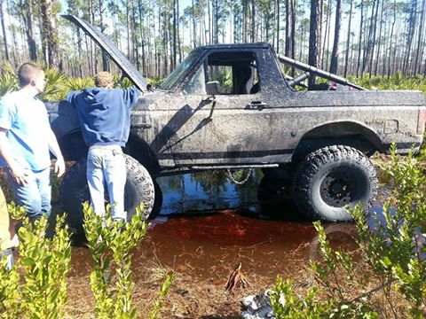 1995 Ford Bronco - A 1995 Ford Bronco is stuck in murky water with its hood up, while two individuals observe the situation in a wooded area.