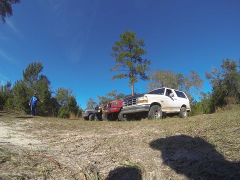 1995 Ford Bronco - Several SUVs, including a 1995 Ford Bronco, are parked on a dirt path under a clear blue sky.