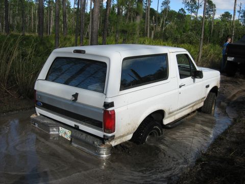 1994 Ford Bronco - A white 1994 Ford Bronco is stuck in the mud in a wooded area, with another vehicle visible in the background.