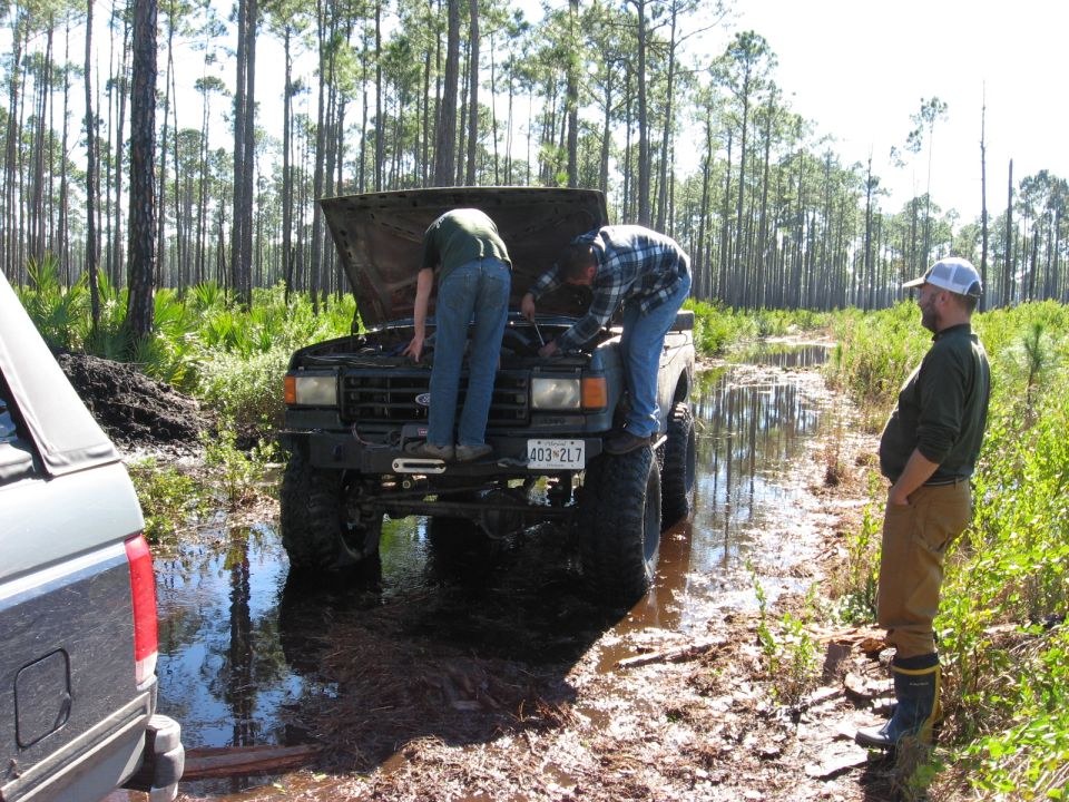Two men work on the engine of a lifted Ford Bronco stuck in mud; another man watches. A second vehicle is partially visible on the left