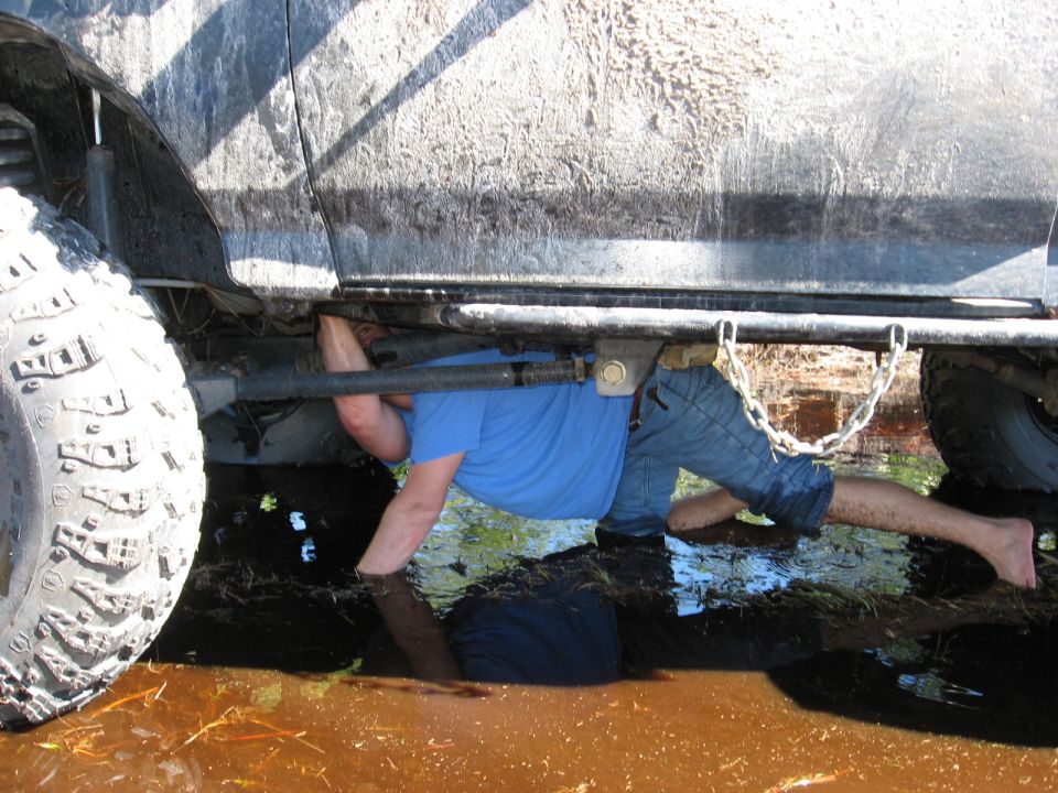 guy working under a muddy SUV in shallow water, big tire visible