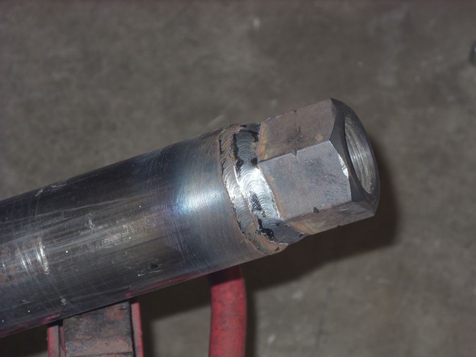Close-up of a metal pipe with a hex nut welded onto the end. The weld shows circular patterns