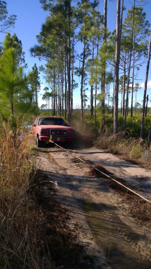 1995 Ford Bronco - A red SUV struggles through a muddy track in a forest, attached to a tow rope.