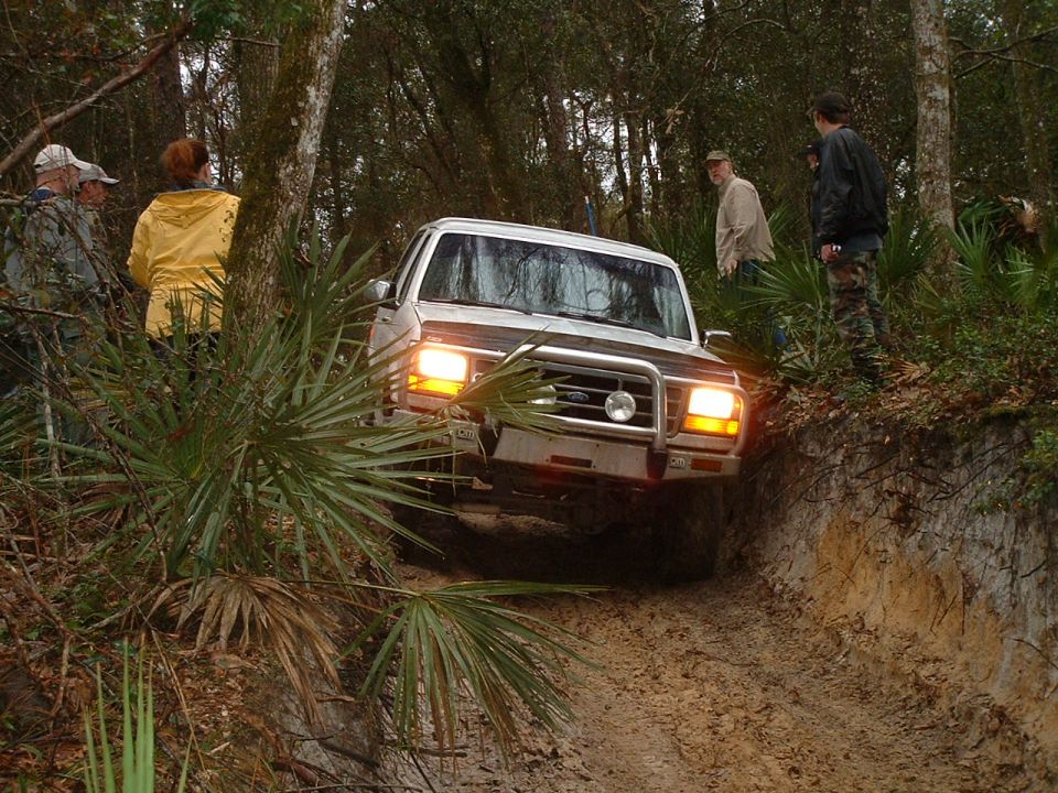 1995 Ford Bronco driving on a muddy trail through trees with people watching