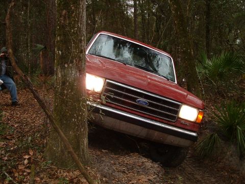 1995 Ford Bronco - A red 1992 Ford Bronco navigates a muddy, wooded trail with its headlights on.