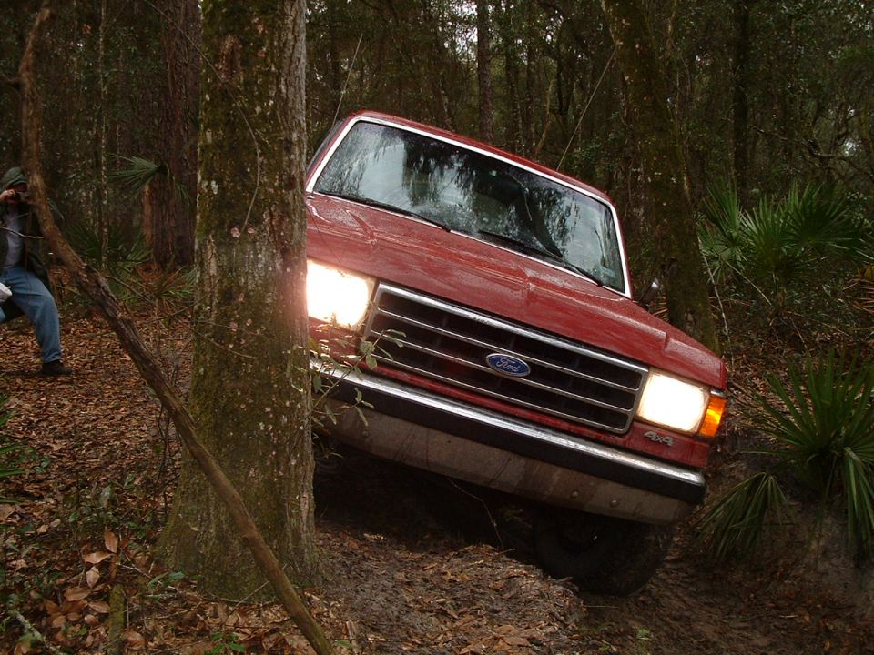 Red 1992 Ford Bronco driving off-road through mud and trees, front-left quarter view