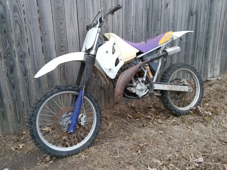 White dirt bike with purple seat and rusty exhaust pipe leaning against a wooden fence