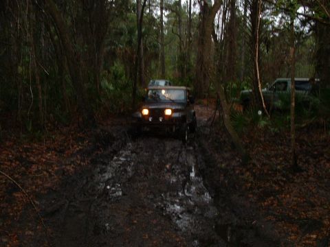1995 Ford Bronco - Two off-road vehicles navigate a challenging muddy trail surrounded by dense trees.