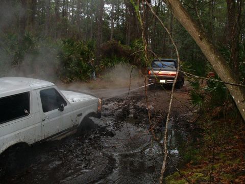 1995 Ford Bronco - A white 1995 Ford Bronco is stuck in the mud while another Bronco drives past on a wooded trail.