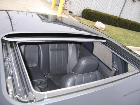 1985 Ford LTD Crown Victoria - View of the rear interior of a car through an open sunroof, revealing black leather seats and cup holders.