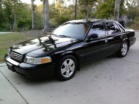 1985 Ford LTD Crown Victoria - A black Ford Crown Victoria four-door sedan photographed from the front-left 3/4 angle, parked on a concrete driveway with grass and trees…