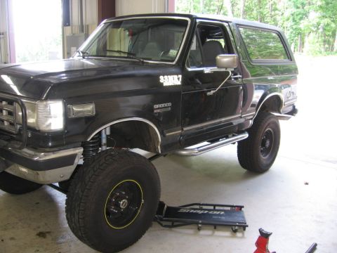 1988 Ford Bronco - A black Ford Bronco is shown with its front wheel raised on a creeper, revealing its suspension and custom wheels.