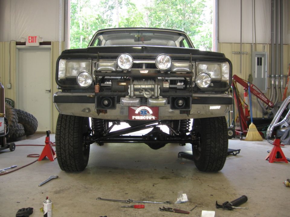 Front view of a modified Ford Bronco, lifted with large tires and a winch mounted on the bumper. The vehicle is in a garage with tools on the floor
