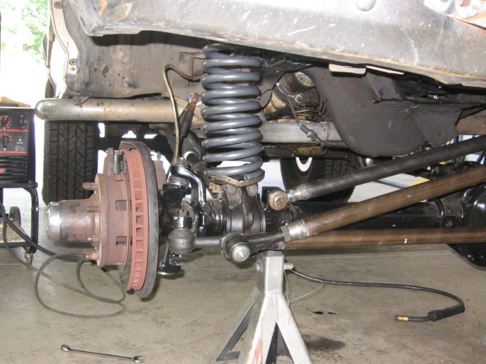 Close-up of a truck's front suspension and brake assembly, with a welding machine in the background