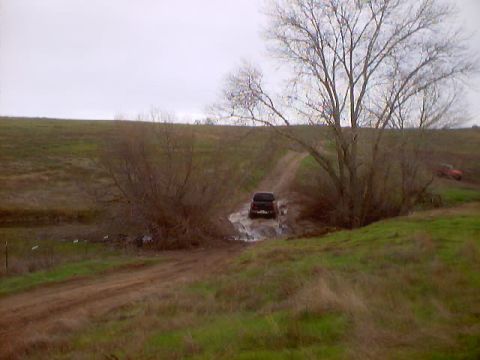 1976 Ford F-100 - A dark SUV navigates a muddy section of a dirt road, with a second vehicle in the distance.