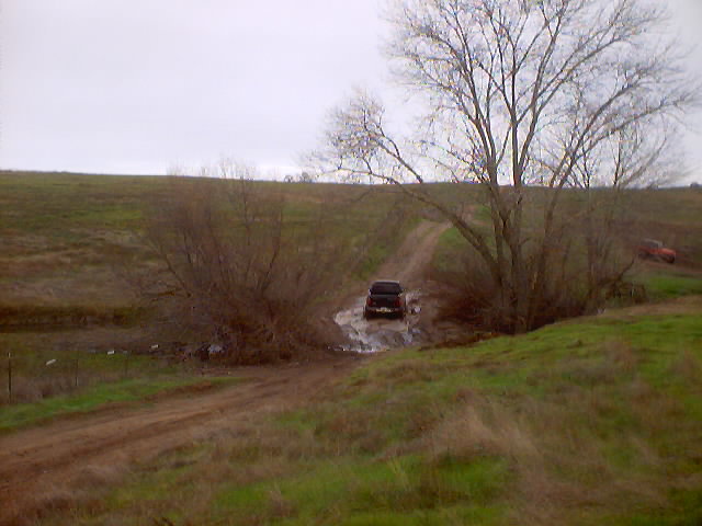 Dark SUV driving through a muddy puddle on a dirt road, with another red vehicle visible on a hill in the background