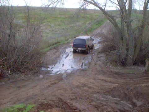1976 Ford F-100 - A blue SUV navigates a muddy track with puddles and tire ruts.