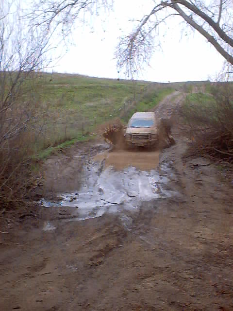 1976 Ford F-100 - A van navigates a muddy obstacle on an off-road trail, kicking up a large spray of water and mud.