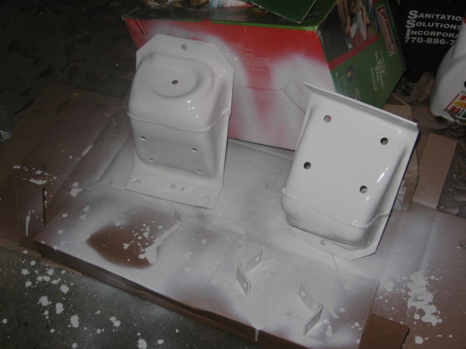 Two white painted metal brackets on a cardboard surface with overspray